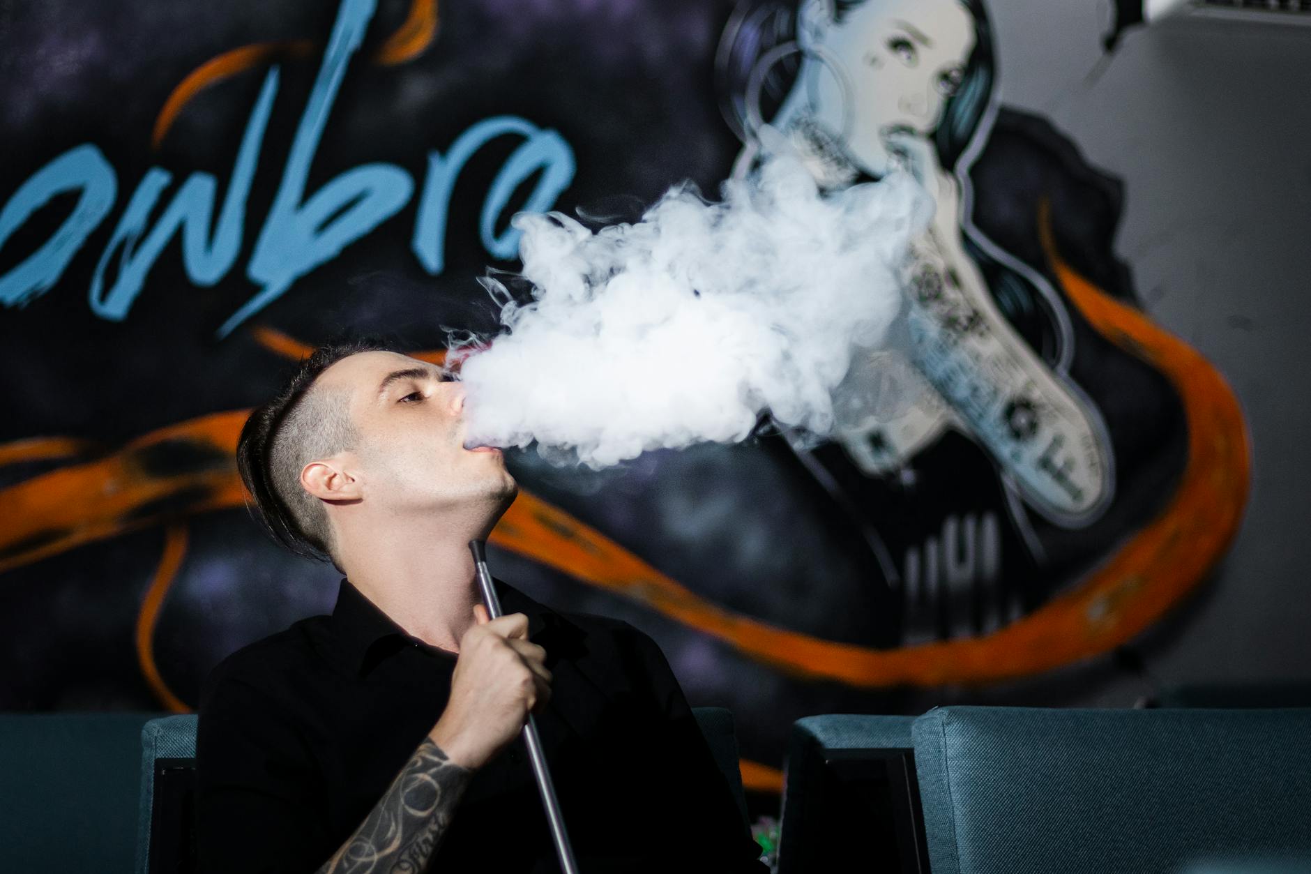 A man with tattoos vapes indoors, creating a smoke cloud against a mural backdrop.